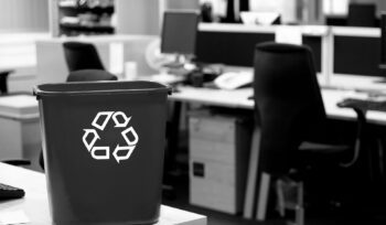 Black recycling bin with white recycle symbol placed on a desk in a modern office with chairs, desks, and computer monitors.