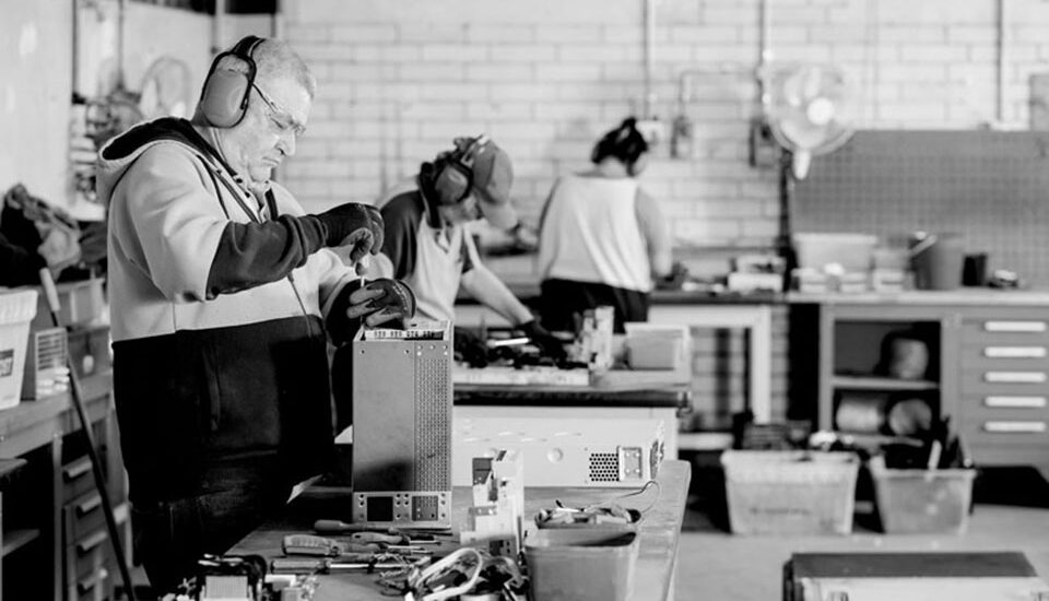 Workers dismantling electronic devices on workbenches with tools and components inside an e-waste recycling facility