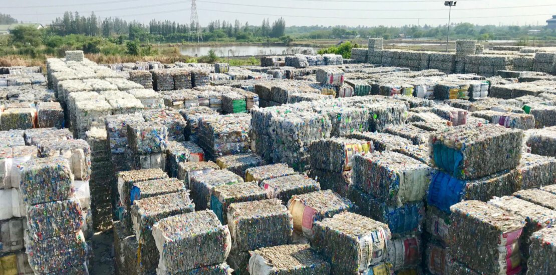 Stacks of compressed plastic waste bales arranged in rows at an outdoor recycling facility, illustrating global waste ban challenges