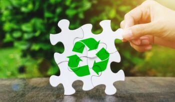 Hand placing a white puzzle piece to complete a green recycling symbol on wood, symbolising plastic recycling and sustainability.