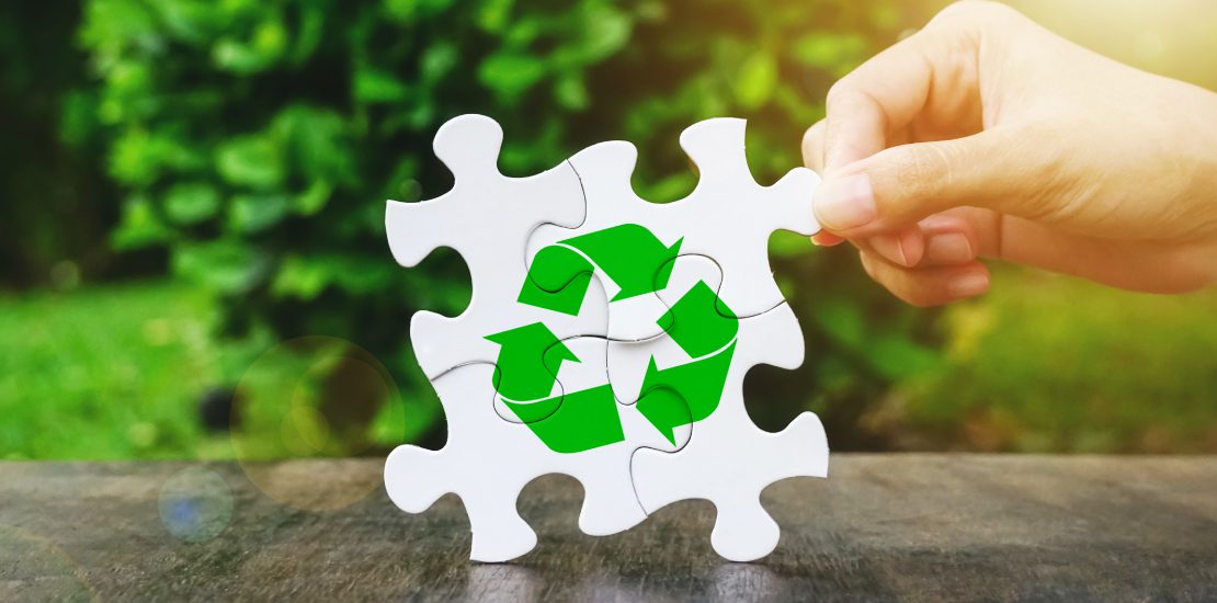 Hand placing a white puzzle piece to complete a green recycling symbol on wood, symbolising plastic recycling and sustainability.