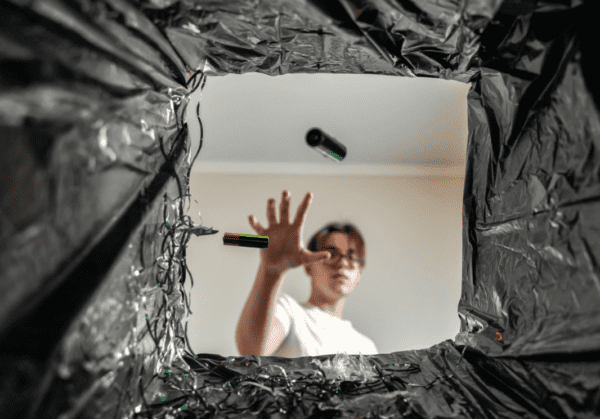 View from inside a black recycling bin looking upward at a person dropping two green AA batteries into the bin, symbolising battery recycling and safe disposal practices.