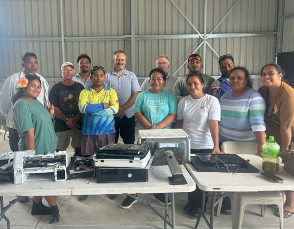 kiribati e waste management training group photo Group photo from Kiribati e-waste training with tables of dismantled printers, computer parts, and electronic components in a workshop.